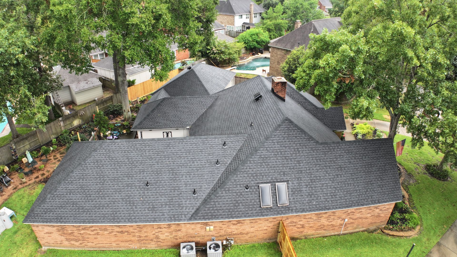 Overhead view of a dark gray shingled roof on a brick house, surrounded by trees and grass.