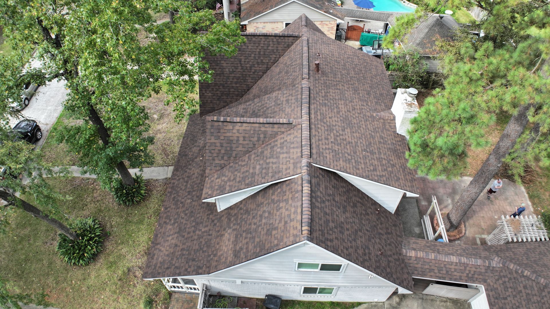 Overhead view of a brown-shingled roof on a white house surrounded by green trees and a backyard