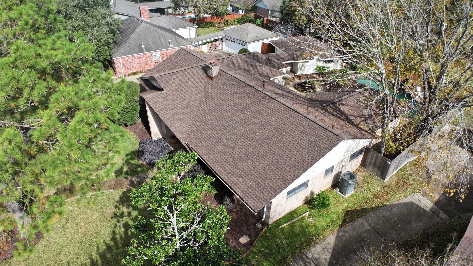 Overhead view of a brick house with a brown roof, surrounded by trees and other houses.