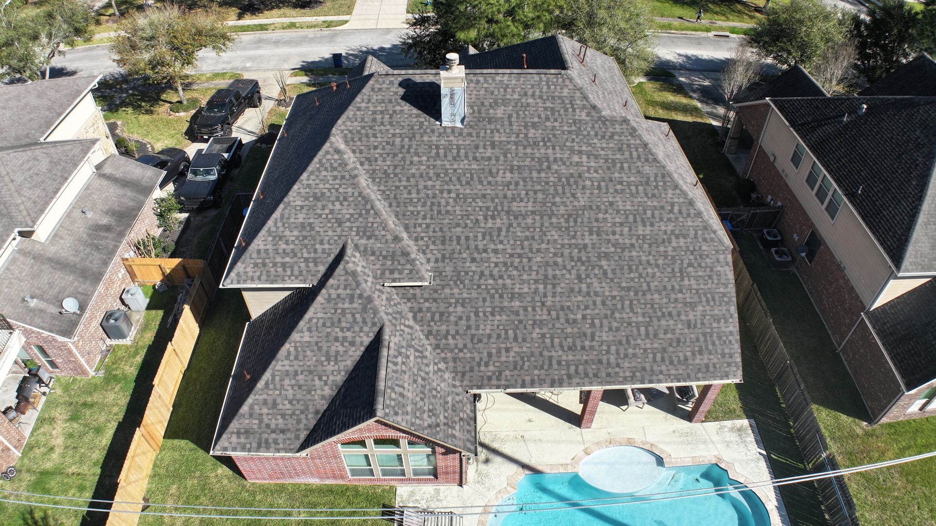 Aerial view of a house with a dark gray roof, chimney, and pool. Adjacent homes and green lawn