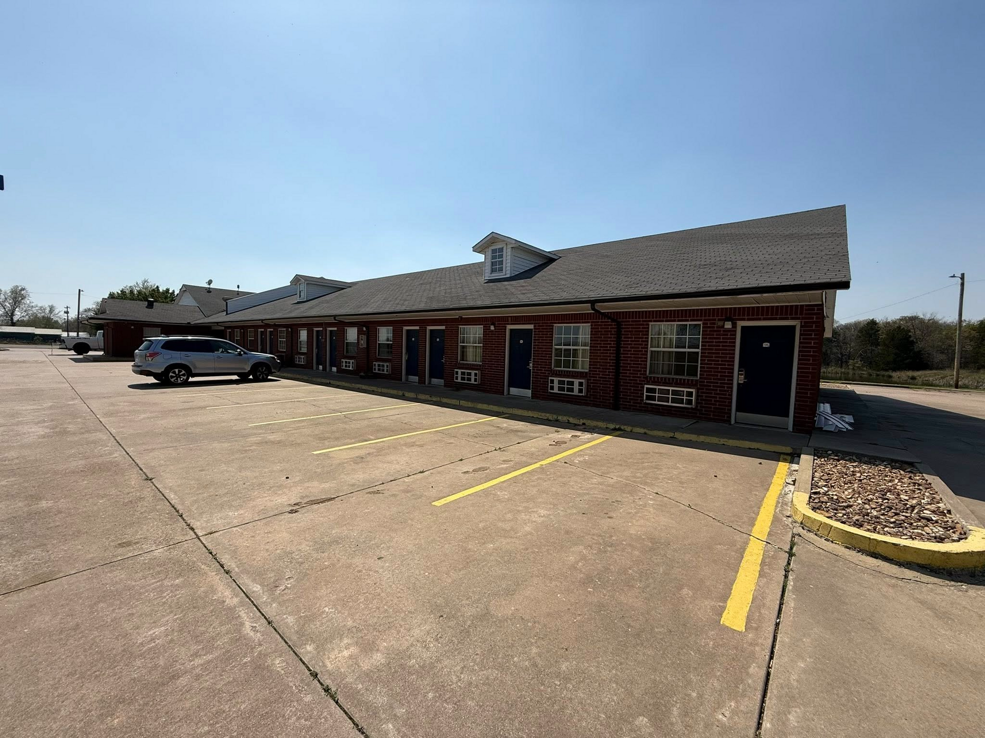 Motel exterior on a sunny day with parking spaces. Red brick building with dark roof and a car.