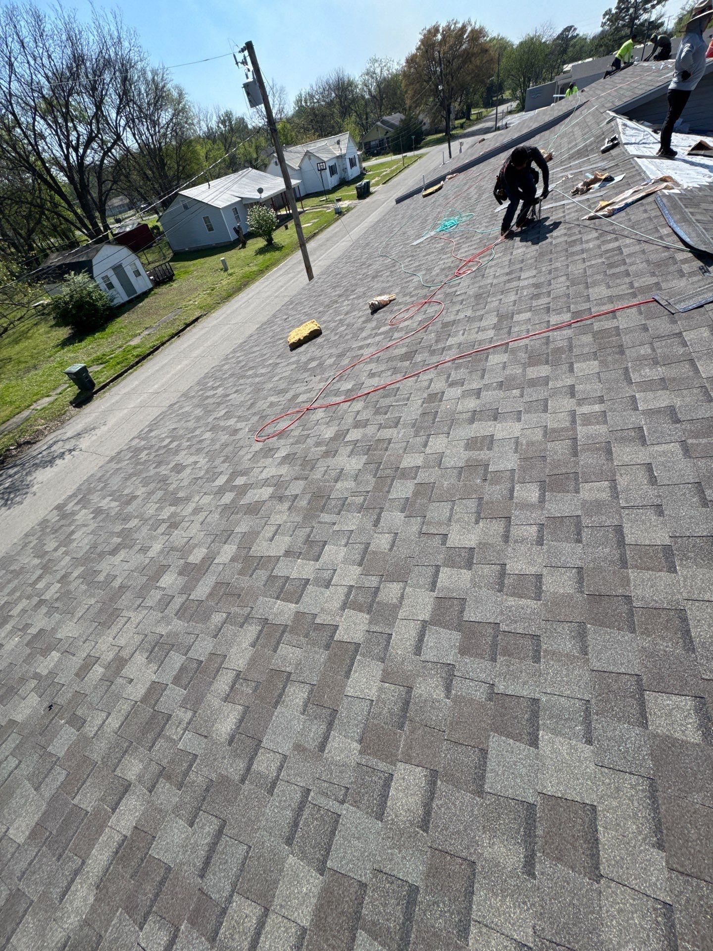 Roofer working on a gray shingle roof. Other roofers and houses in the distance. Blue sky and sunny.