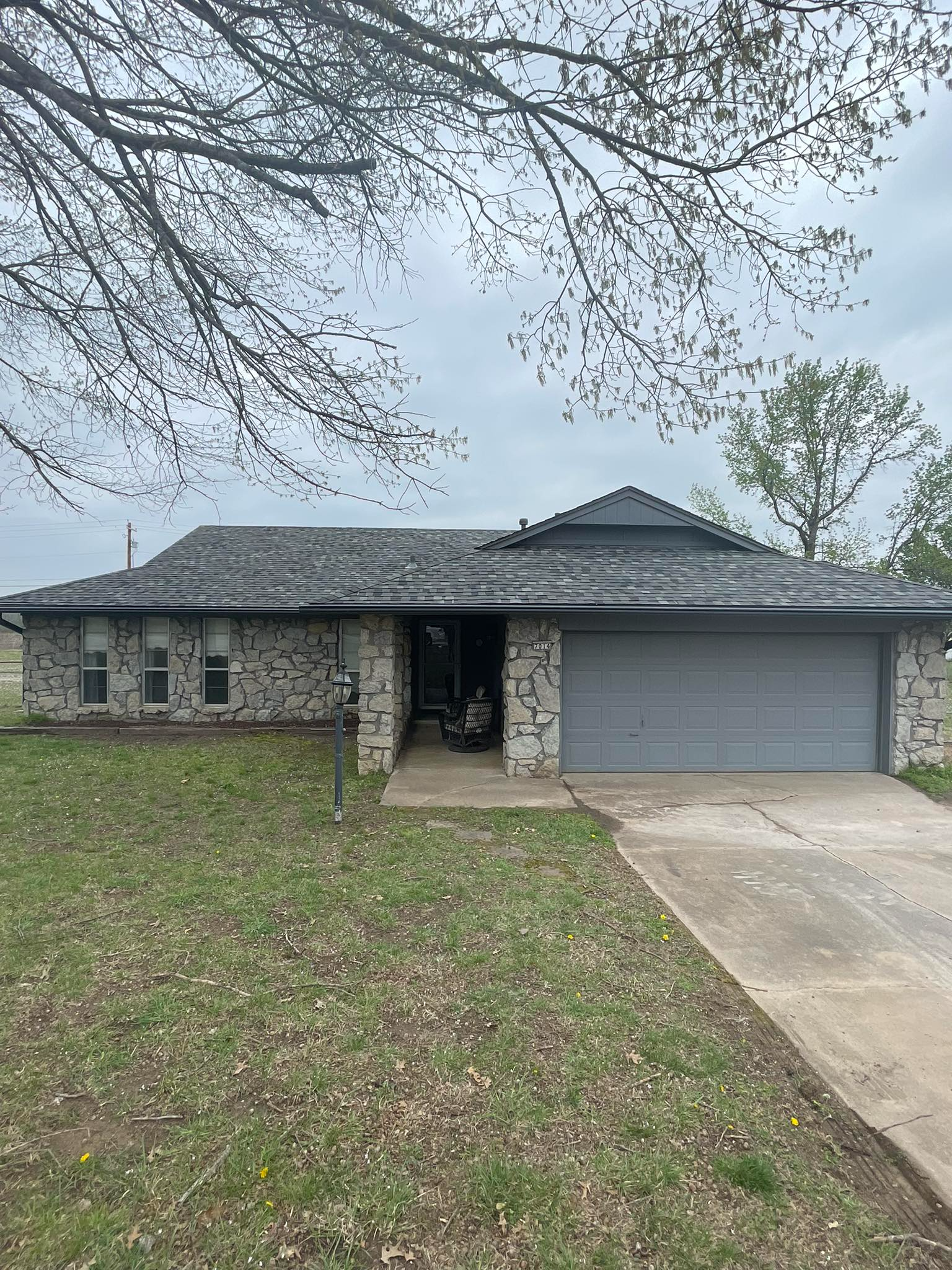 Gray-roofed ranch house with stone exterior, overgrown grass, cloudy sky.