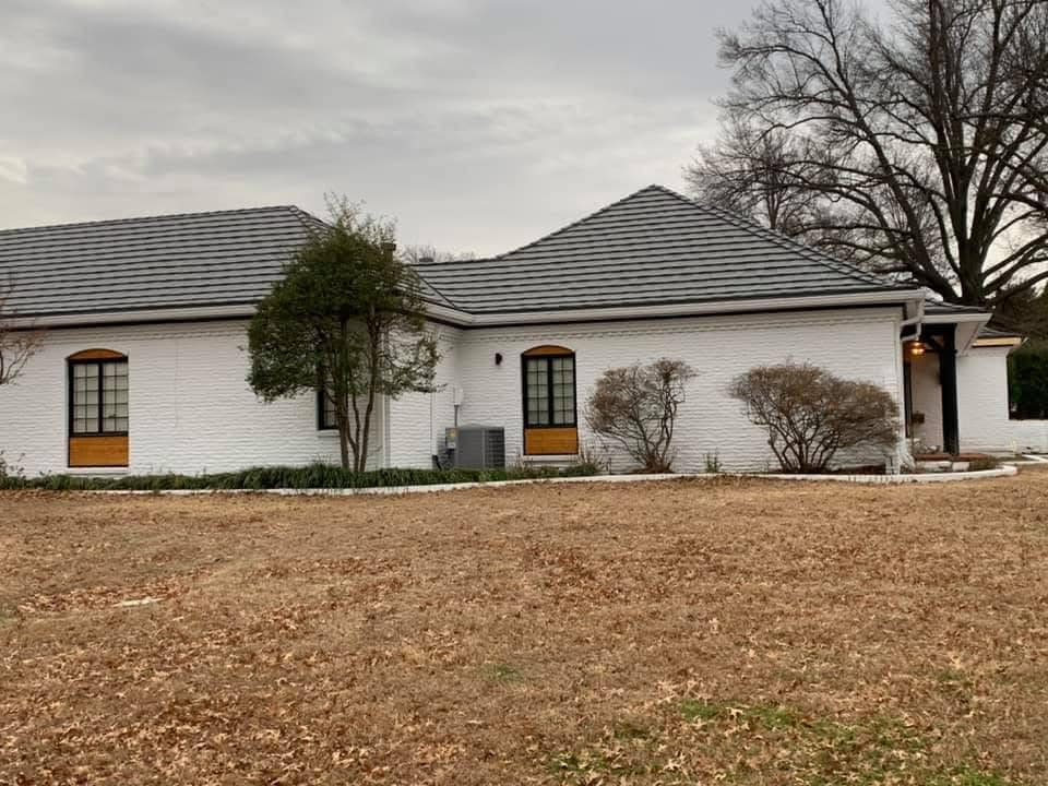White brick house with gray roof, windows, and overgrown landscaping on a cloudy day.