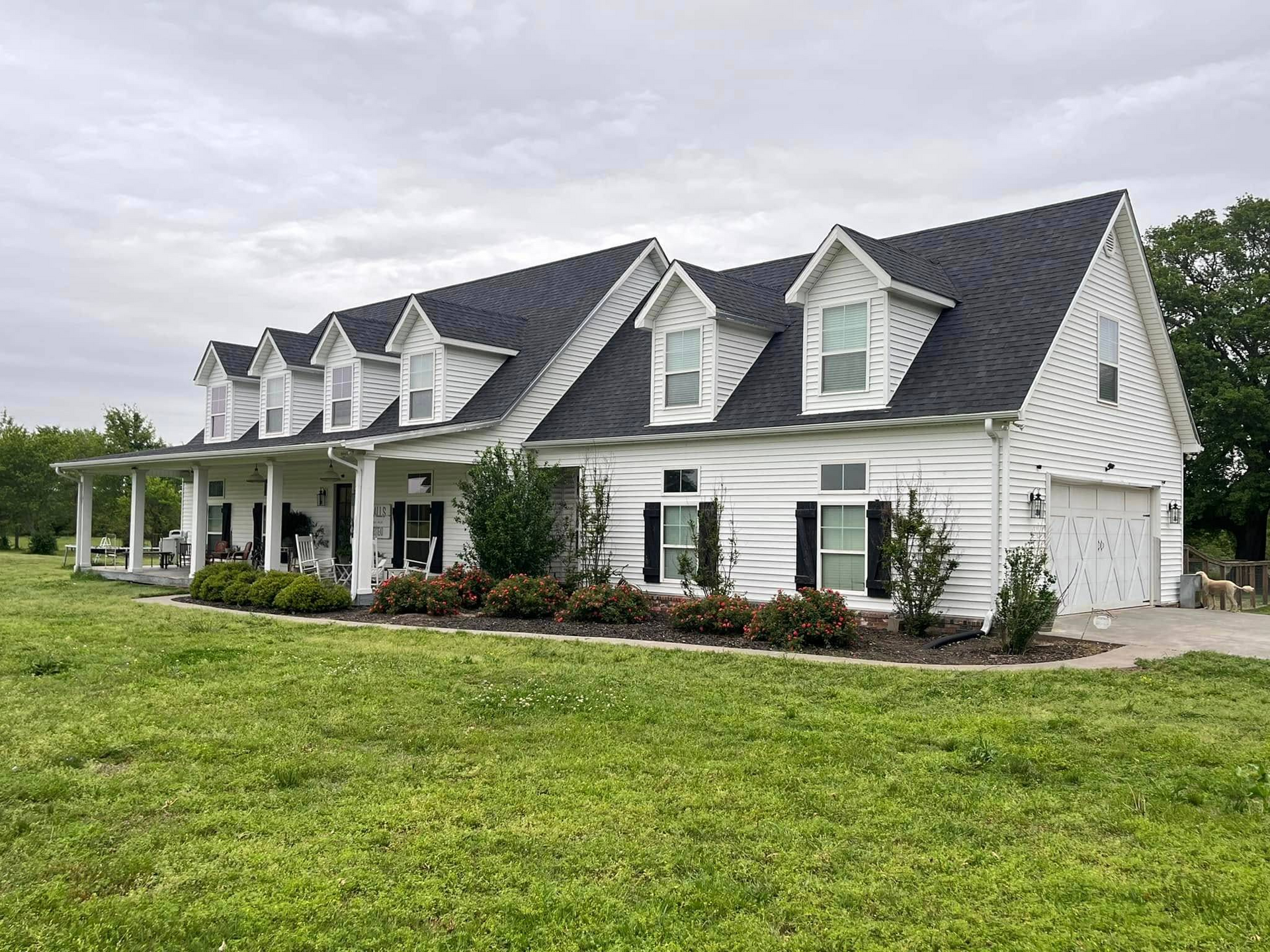 White house with black shutters, dormers, and a long front porch; green lawn.