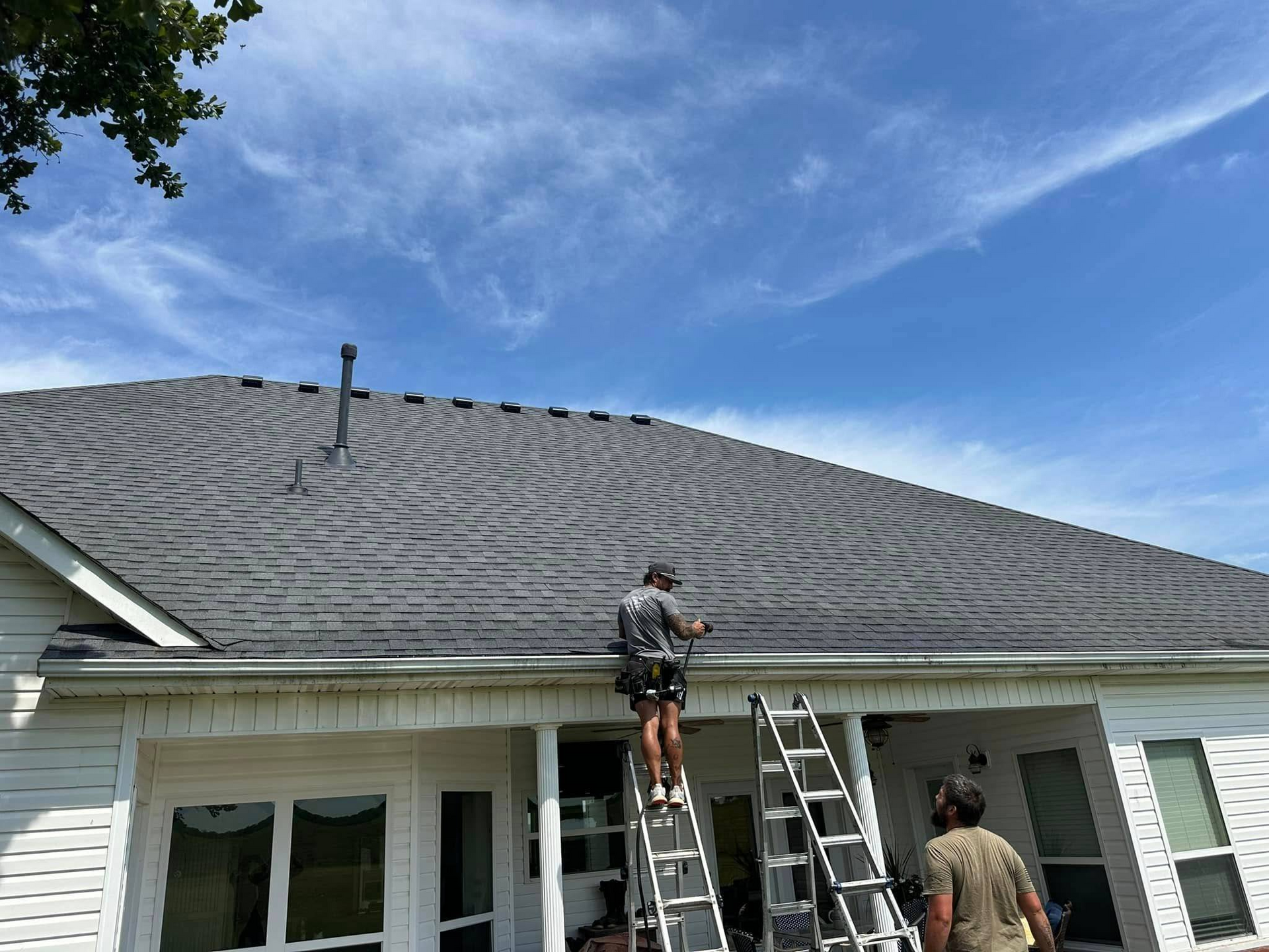 Two workers on a roof, one on a ladder. House has dark roof, white siding, and blue sky.