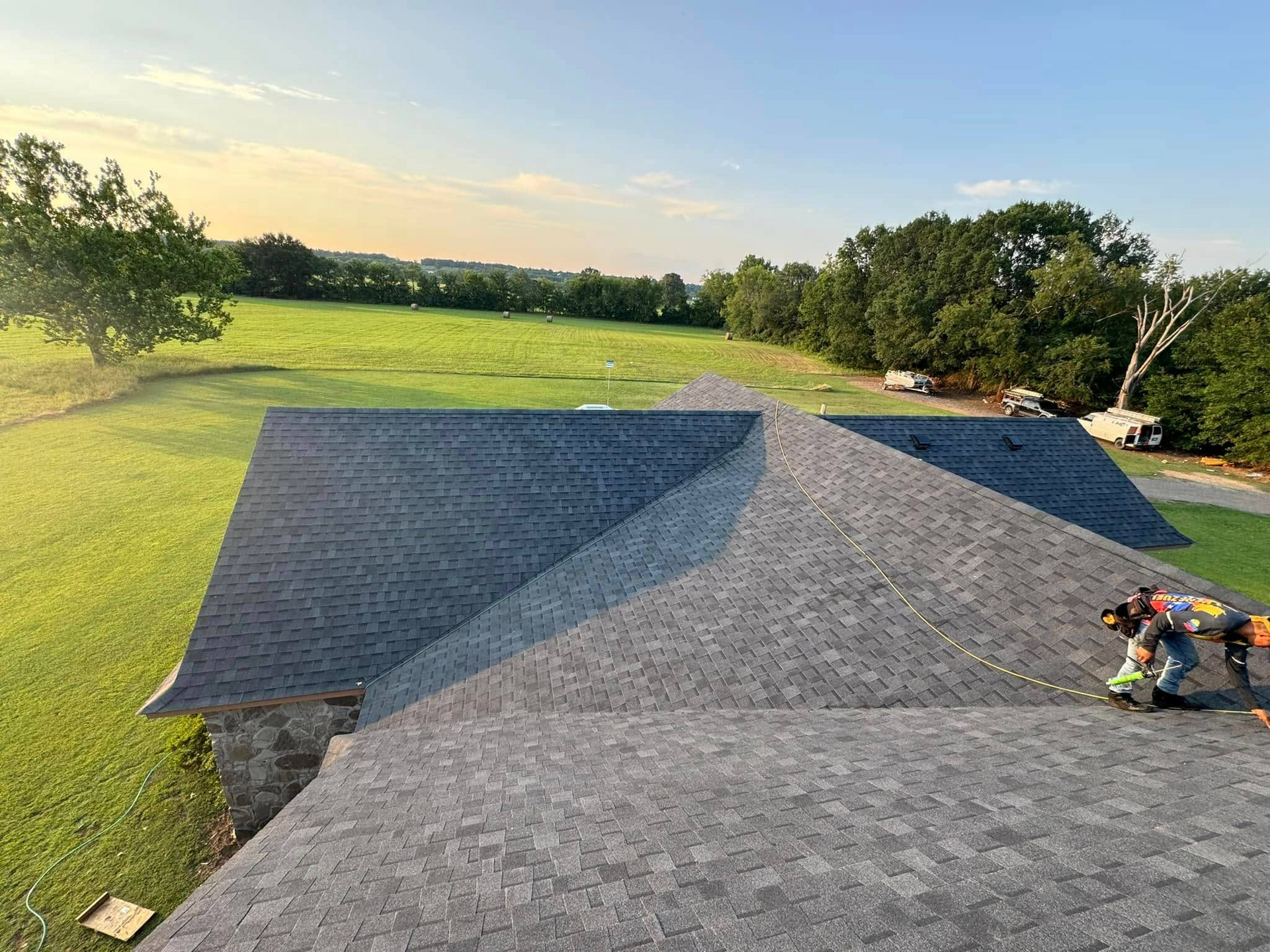 Roofers working on a gray shingle roof of a house, set in a green field with trees under a blue sky.