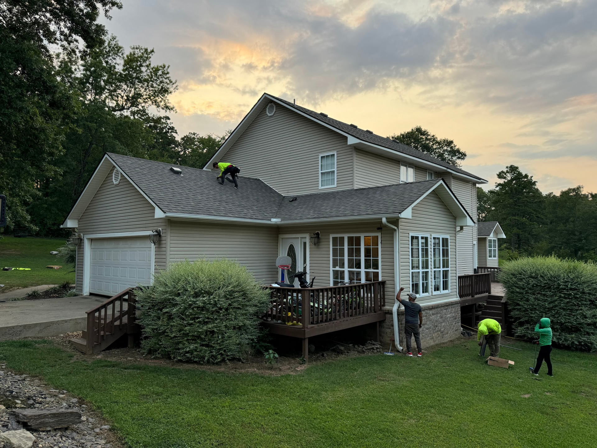 Workers on a roof, near a house with a garage and deck, and workers on the ground. Green grass and trees are present.