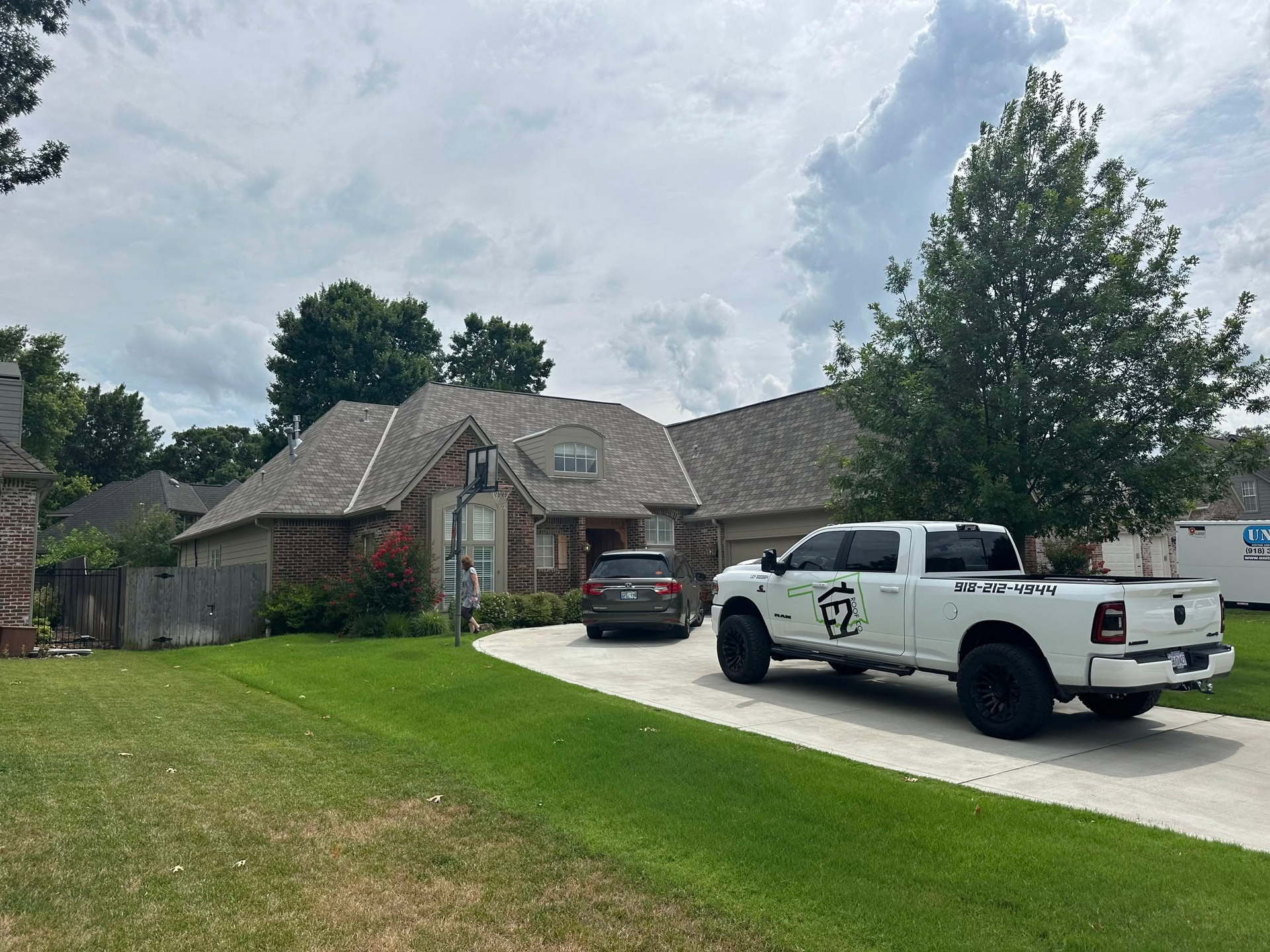 A brick house with a white truck parked in the driveway and a gray car. Green lawn, trees, and cloudy sky.