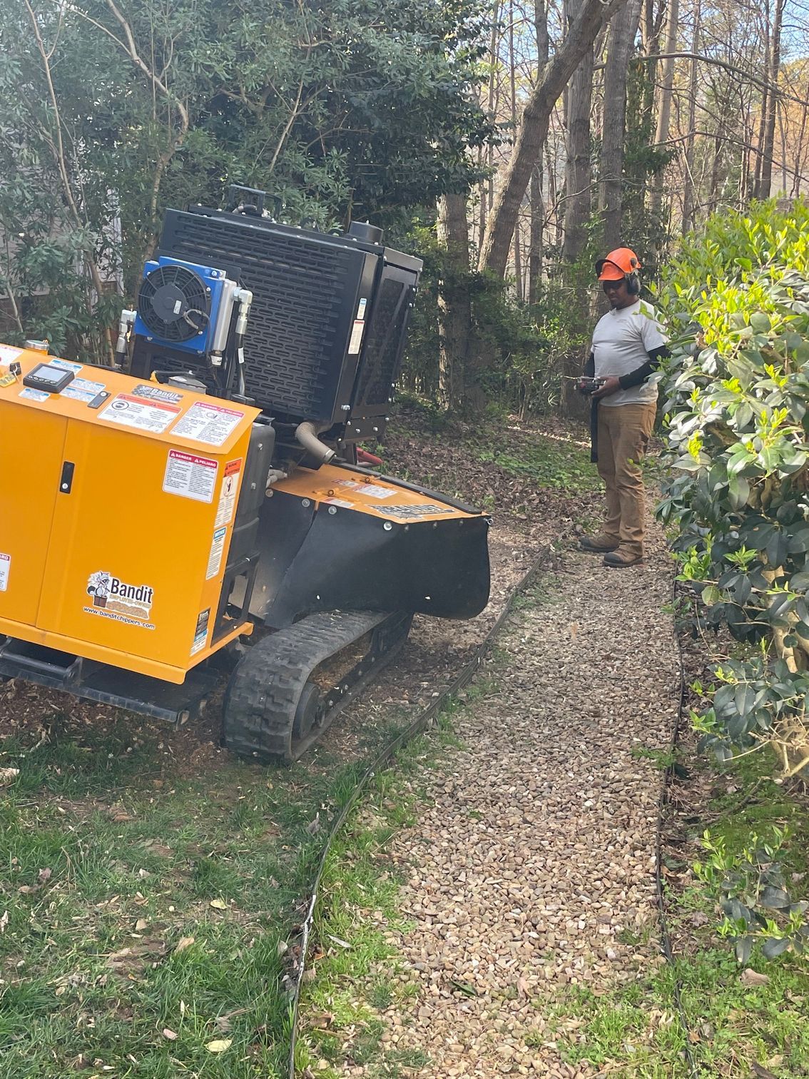 Orange construction machine on a narrow wooded path with a worker in an orange hard hat nearby.