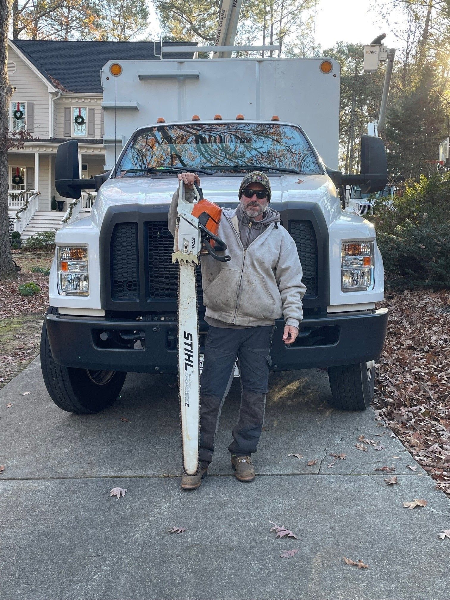 Person standing in front of a white truck, holding a long white pole with an orange tool attached.