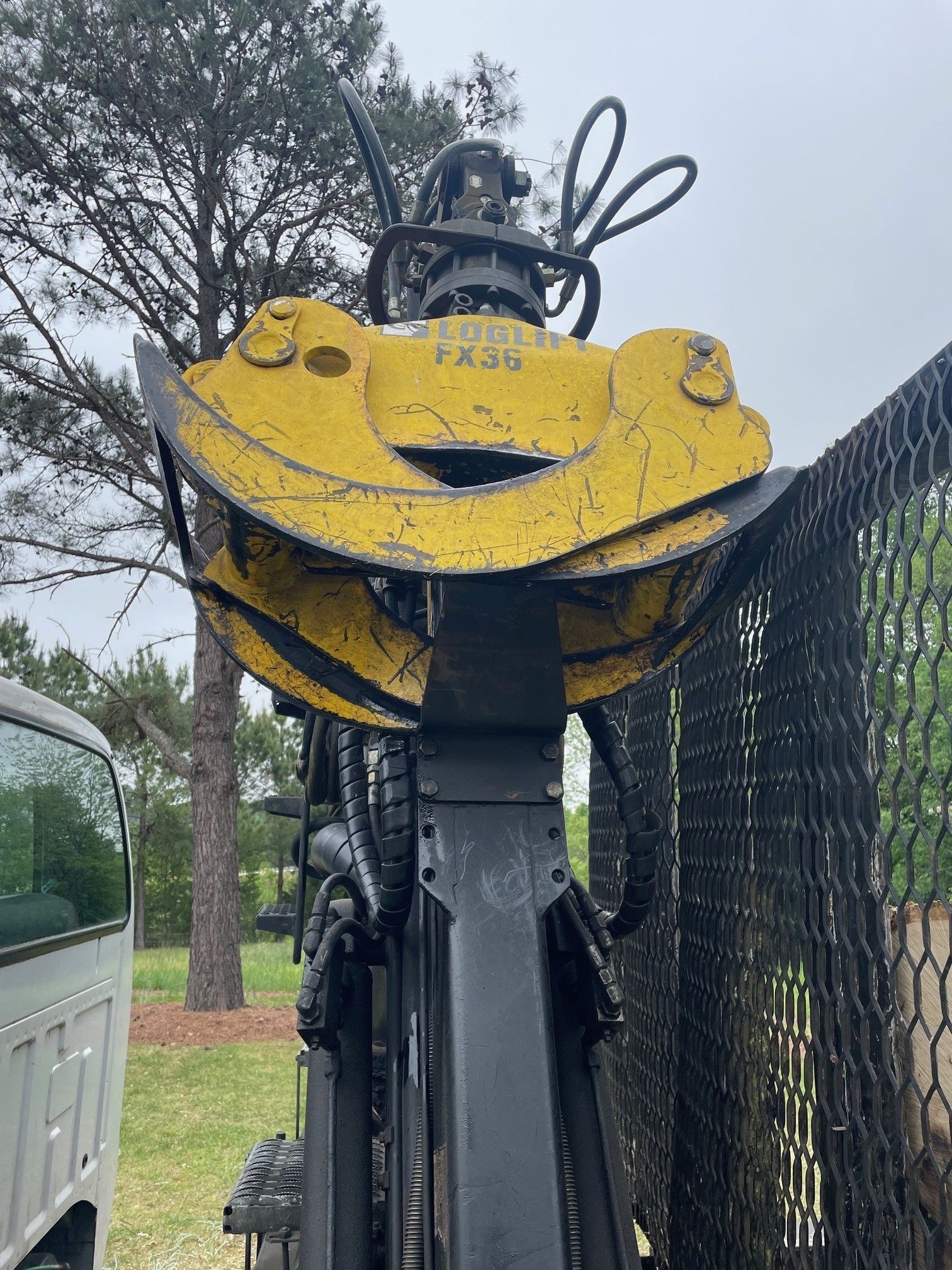 Yellow industrial machine arm beside a fence, with a truck and trees in the background