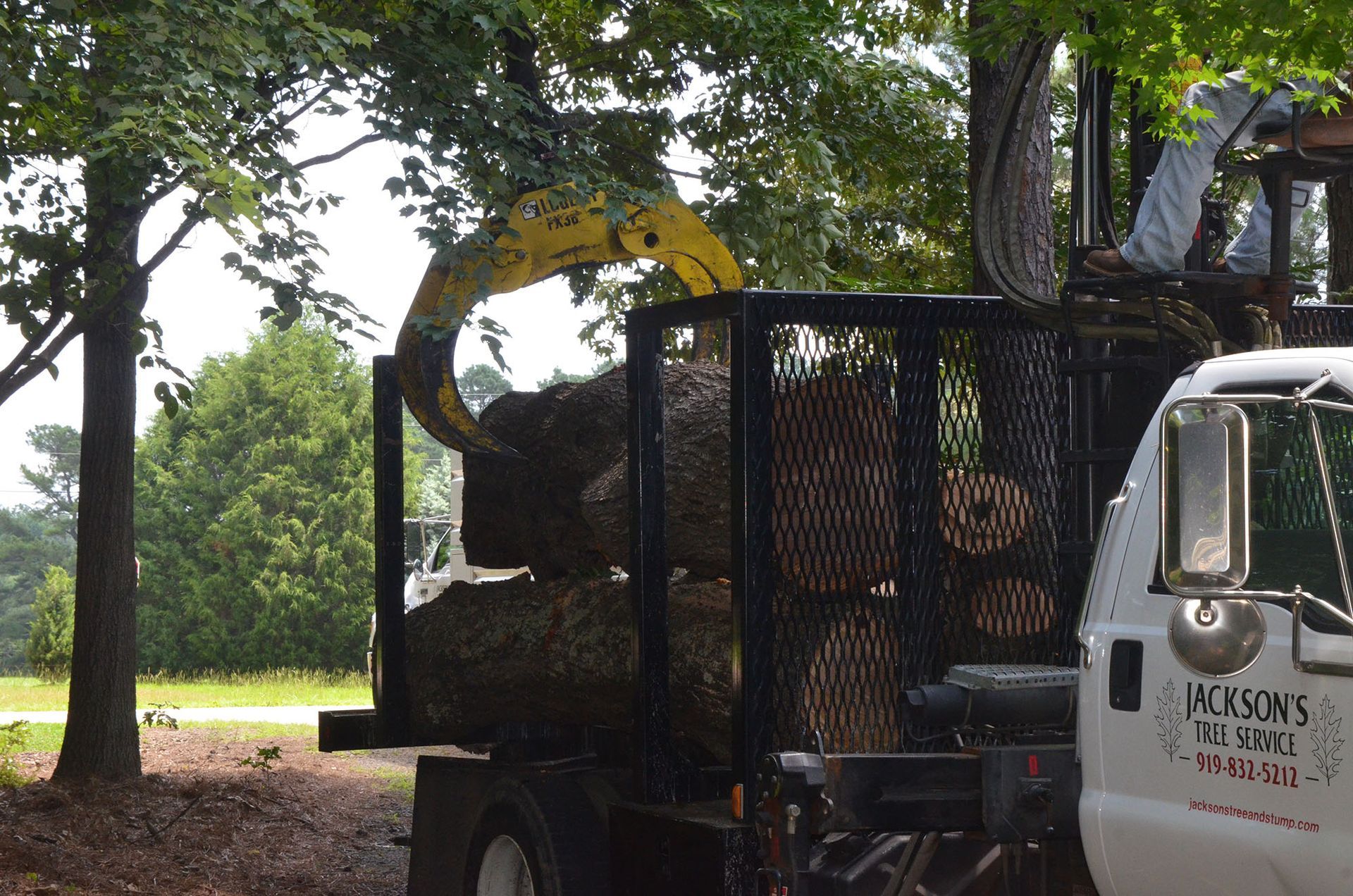Logging truck loading cut branches in a wooded area with a yellow excavator arm.