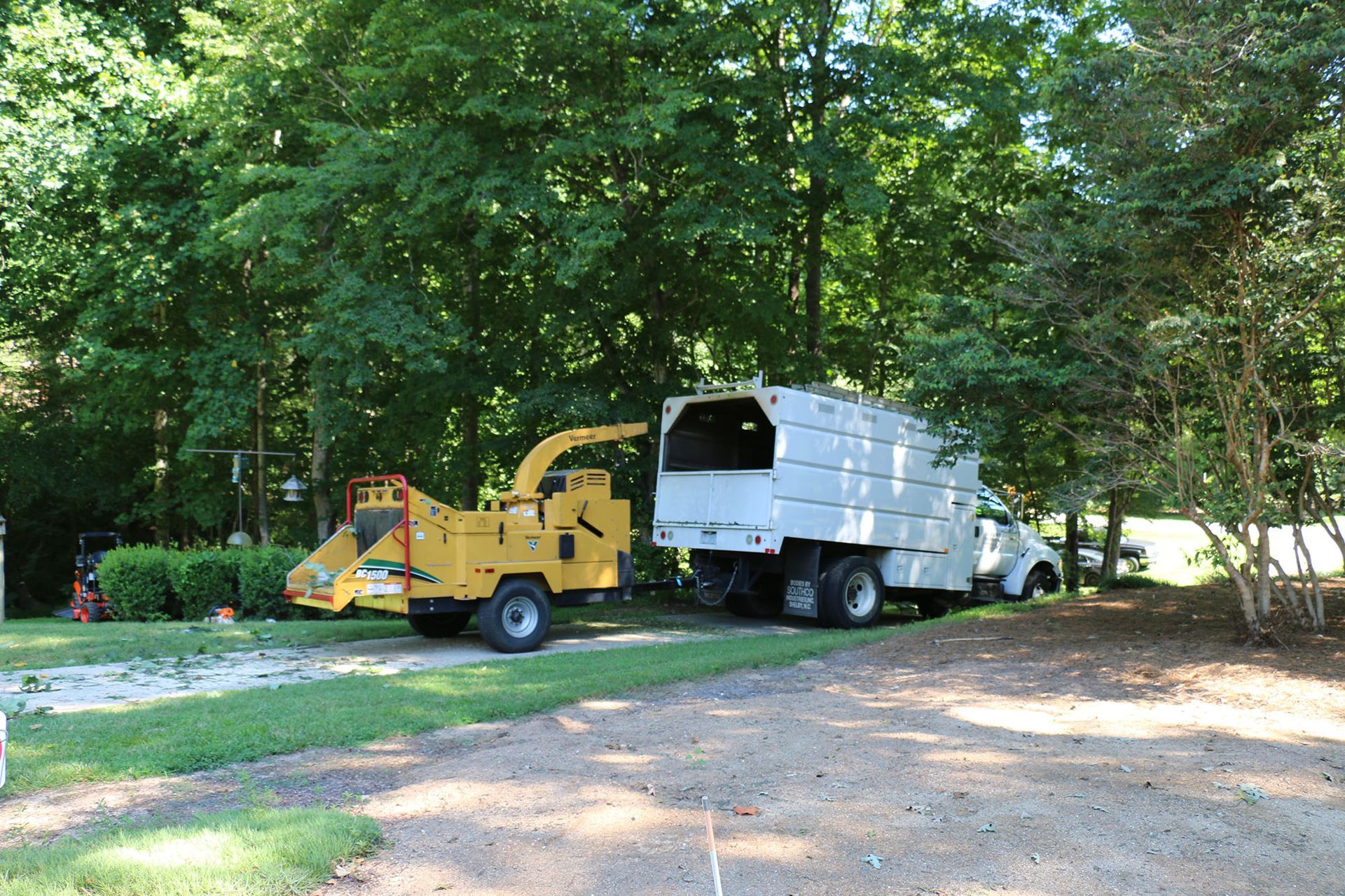 Yellow wood chipper and white truck on a wooded driveway, with logs and trees nearby