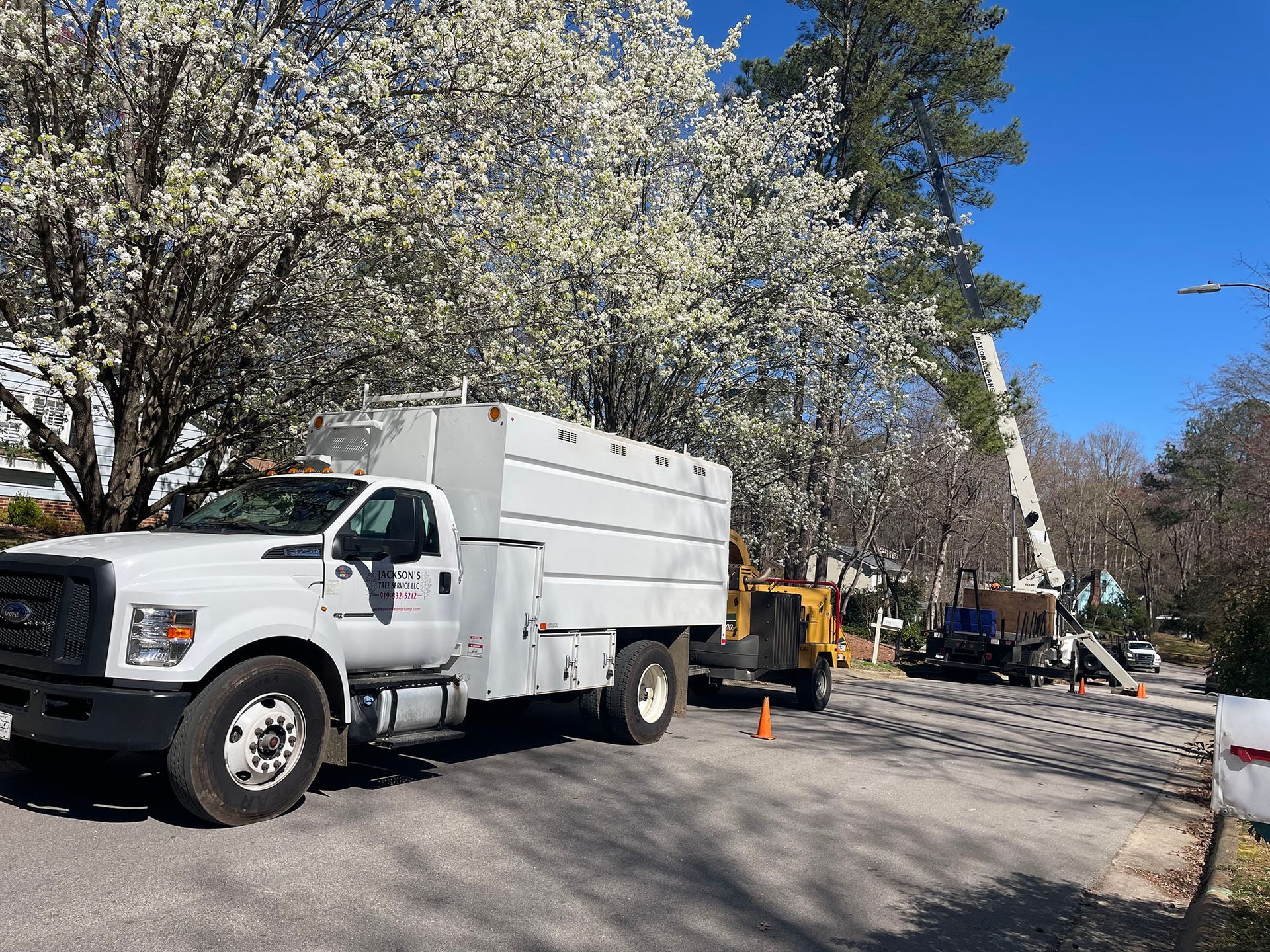 White utility truck and construction equipment on a tree-lined street under blooming trees.