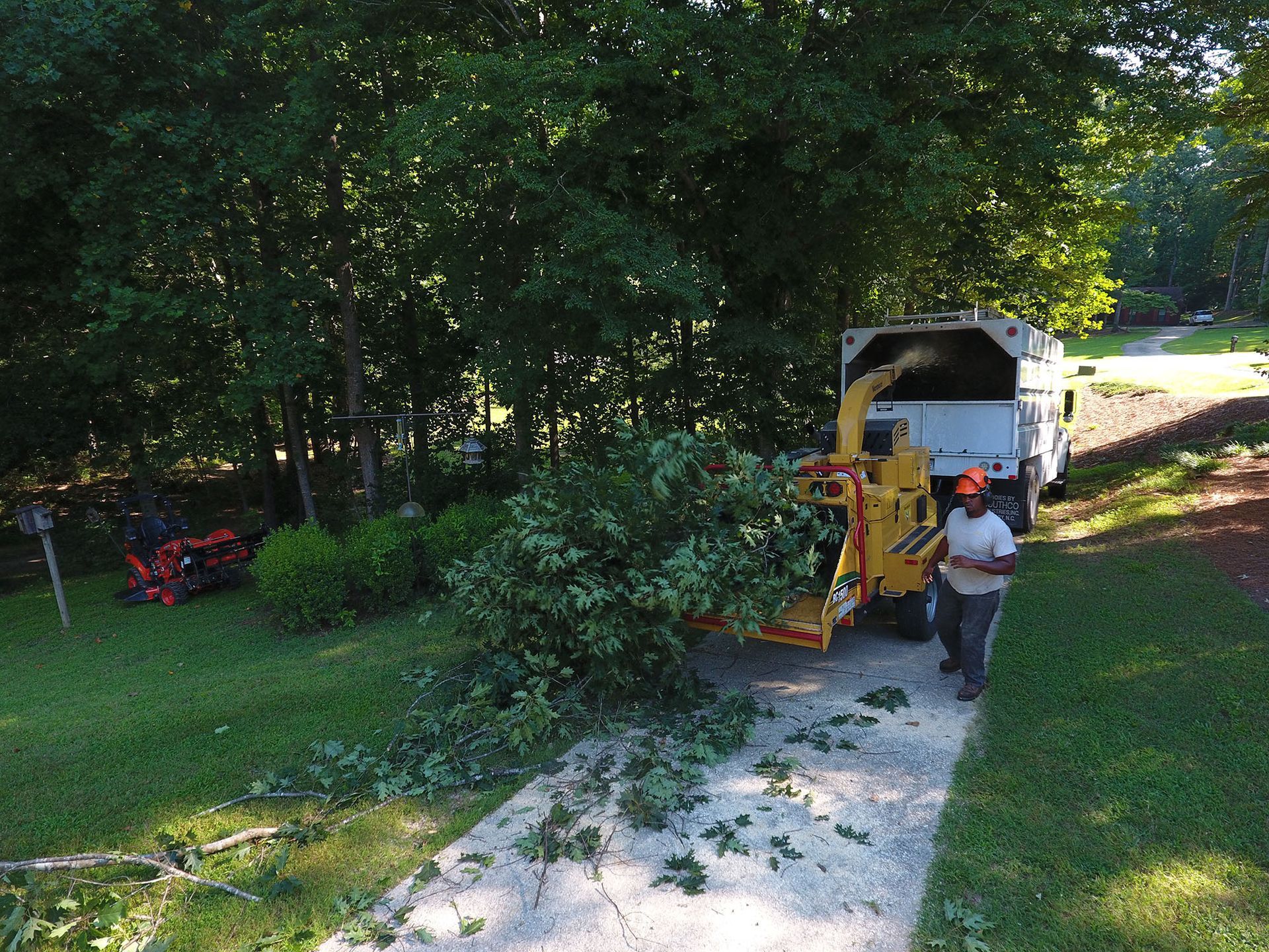 Worker feeding tree branches into a wood chipper beside a white truck on a wooded driveway