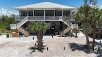Elevated white beach house with screened porch, two staircases, on sandy ground, palms.
