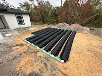 Black septic drain field tubes arranged on sandy ground near a house under construction.