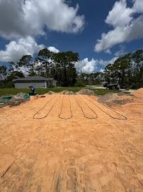 Sandy construction site with pre-laid foundation outlines. Bright sky, trees, and houses in the background.