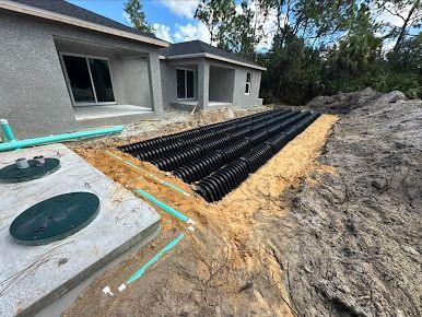 Construction site with septic tank and drain field near a house. Dark corrugated pipes in a trench.