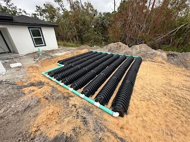 Installation of a septic drain field with black corrugated pipes and green PVC pipes on a sandy lot next to a house.