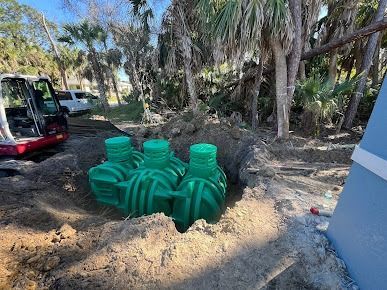 Green septic tanks in a hole, an excavator, trees, and a light blue wall.