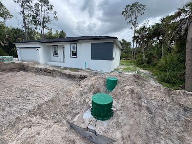 A white house under construction with a septic system in the foreground. Green tanks, dirt, and trees are visible.
