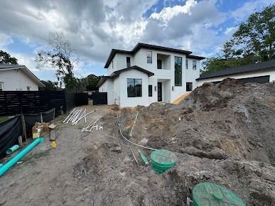 New white two-story house under construction, dirt pile in front, pipes, and black fence.