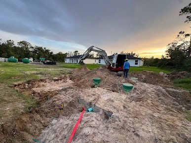 Construction site with excavator and person, trenches, green septic tanks, and house in background.