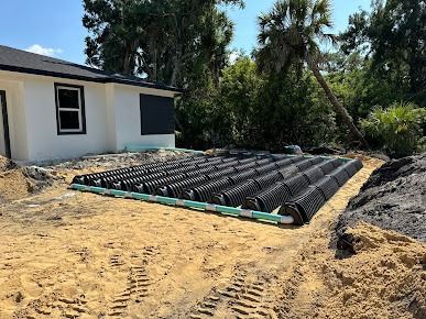 Black septic system components beside a house, surrounded by sand and dirt.