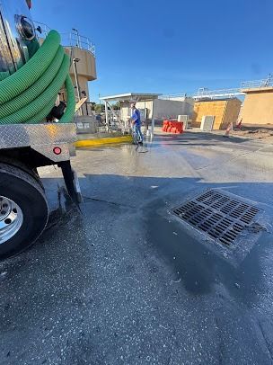 A worker sprays a paved area near a sanitation truck and facility.