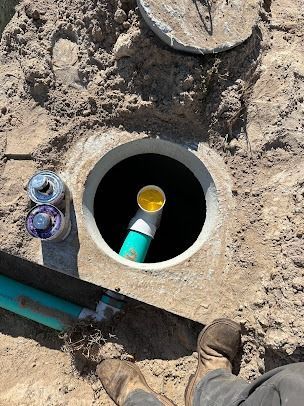 Open manhole in dirt. A person's boots are visible. PVC pipes and concrete lid are nearby.