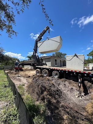 A crane lifting a large rectangular container off a flatbed truck near a small house under construction.