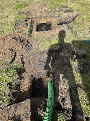 Hole dug in grass; green hose extends into it; shadow of person with shovel visible.