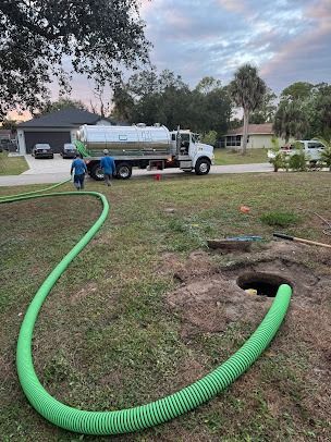 Septic truck parked on a lawn, green hose connected to an open hole. Two people walk.