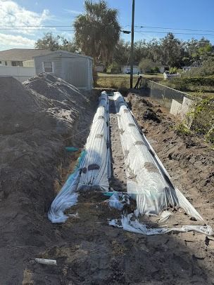 Trench with two covered pipes, likely for utilities, in a dirt yard near a small shed and fence.