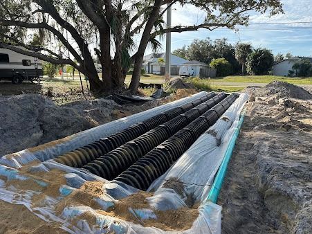 Installation of a drain field system: black corrugated pipes in a trench, surrounded by sand and fabric. Outdoor setting.
