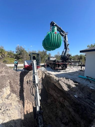 An excavator lifts a large, green septic tank over a trench at a construction site.