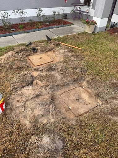 Two square septic tank covers are exposed in a dug-up lawn. Shovel and paint can nearby.