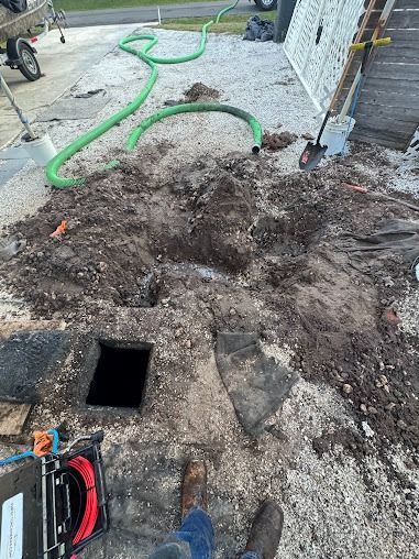 Hole in ground, dirt pile, and green hose on gravel. Person wearing boots stands nearby, tools visible.