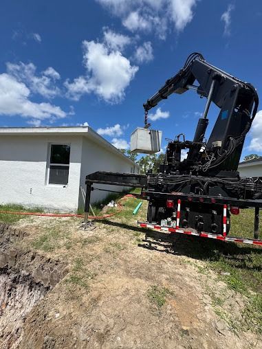 A crane lifting a large, square object near a white house with a blue sky background.