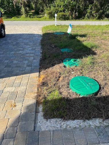 Green septic tank covers in a patch of grass next to a brick driveway.