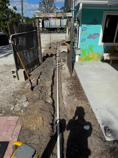 Trench with white pipe, alongside building with mural. Person's shadow visible.