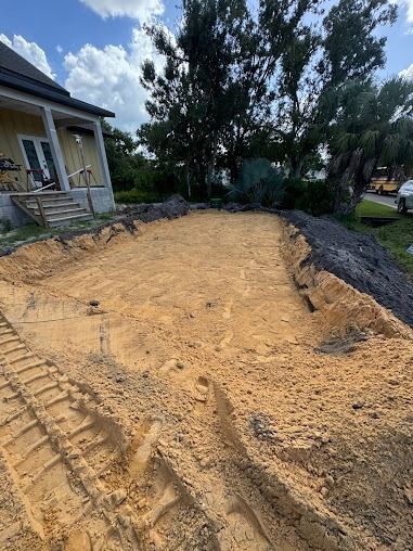 Construction site: leveled dirt area next to a house with a pile of dark material, sunny day.