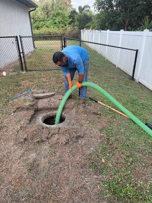 Man in blue shirt, using green hose to service a septic tank in a grassy yard.