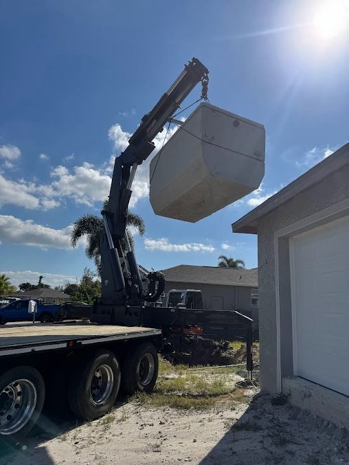 A crane lifting a large, light-colored object over a residential building on a sunny day.