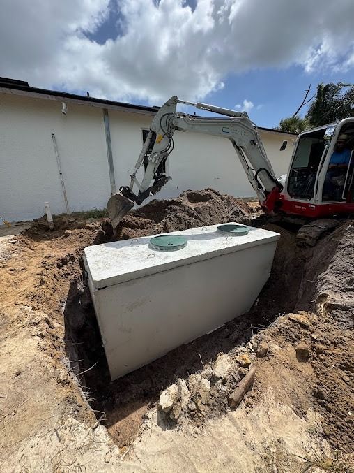 Excavator placing a concrete septic tank in a deep hole near a building on a sunny day.