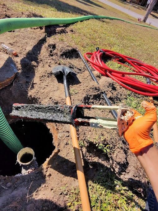 Manhole excavation with tools: shovel, rake, and hose. Orange-gloved hand holds piece of pipe. Green and red hoses.