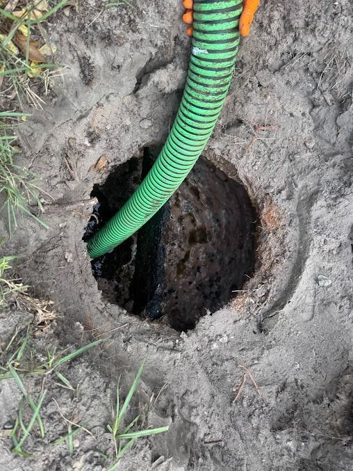 Hole in the ground with a green hose, possibly for pumping waste.  Surrounding dirt and grass.