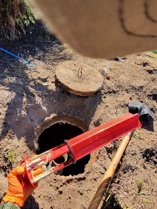 Person holding a red tool over an open manhole. Orange-gloved hand. Ground and cover visible.